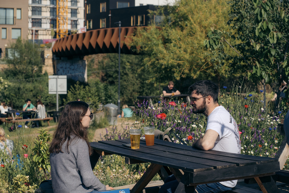 A man and a woman sit at a picnic table in a garden by the river Aire. In the background is a footbridge and flats.