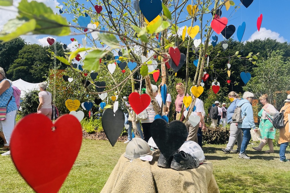 Brightly coloured wooden heart shapes hang from a tree, a table covered in hessian with hard hats and work boots on it. Members of the public make poignant tributes in marker pen to lost loved ones on the colourful wooden hearts at the Constructing Minds RHS show garden.