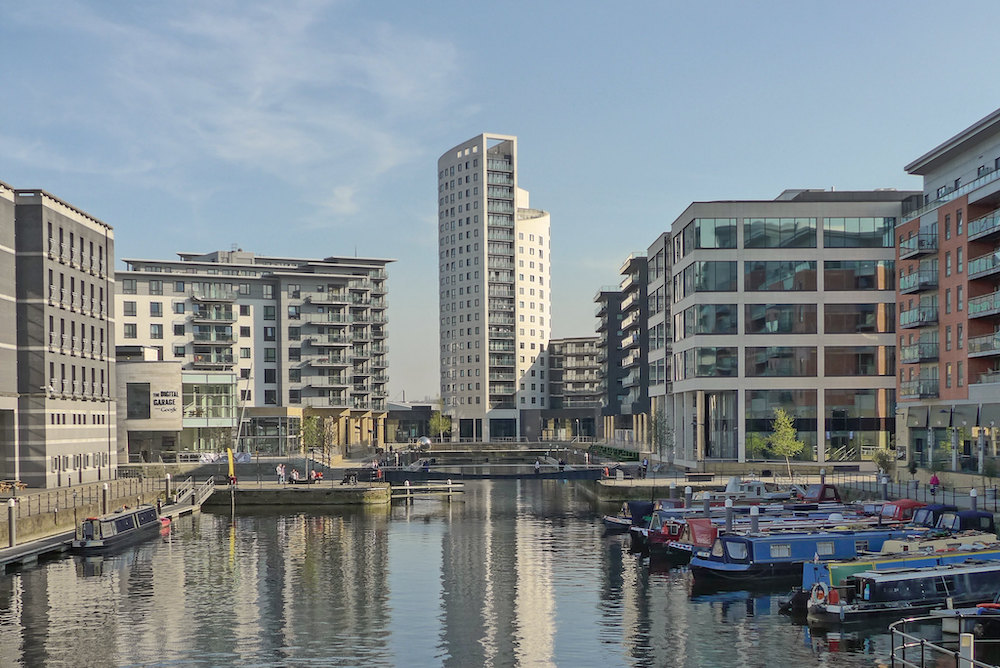 A view looking down Leeds Dock. A cental tower block and lower rise apartment blocks on each side are reflected in the still water of the dock. There are barges moored to the right hand side.