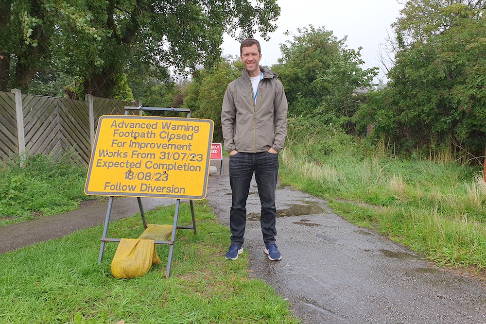 Cllr Andrew Scopes stands next to a sign saying works to improve the paths behind Asda on Old Lane in Beeston will take place between 31/07/23 - 17/08/23